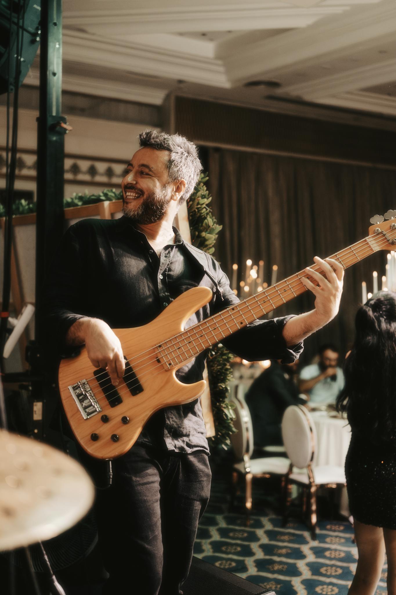 Smiling bassist performing energetically at a lively indoor party setting, surrounded by elegant decor.